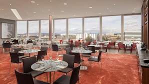 A spacious dining area with multiple tables and chairs, large windows offering a city view, and a modern ceiling design. - Hilton Zurich Airport (Opfikon)