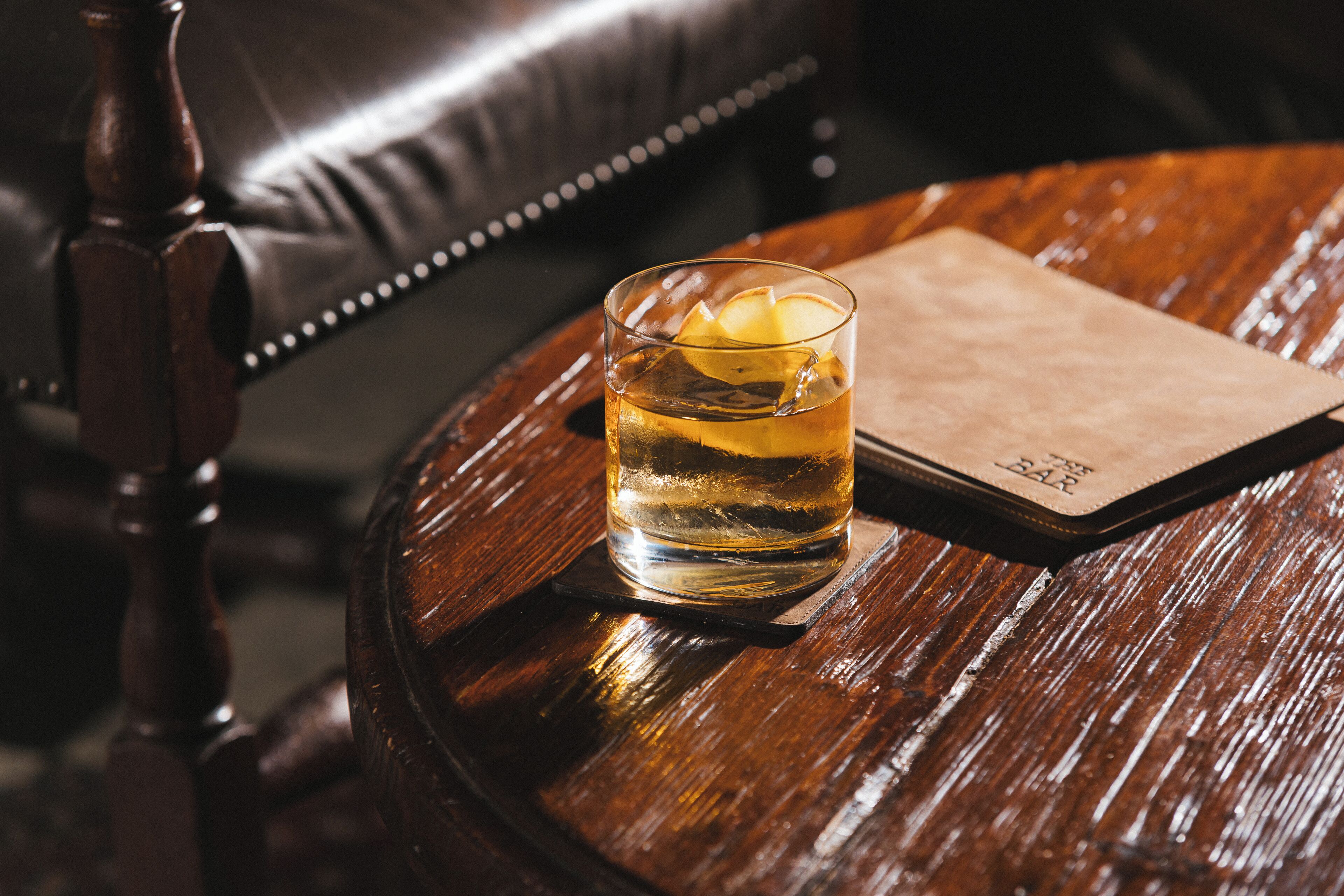 A glass of amber liquid with a lemon slice on a coaster, placed on a round wooden table with a leather chair in the background.