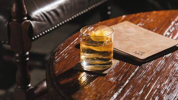 A glass of amber liquid with a lemon slice on a coaster, placed on a round wooden table with a leather chair in the background.
