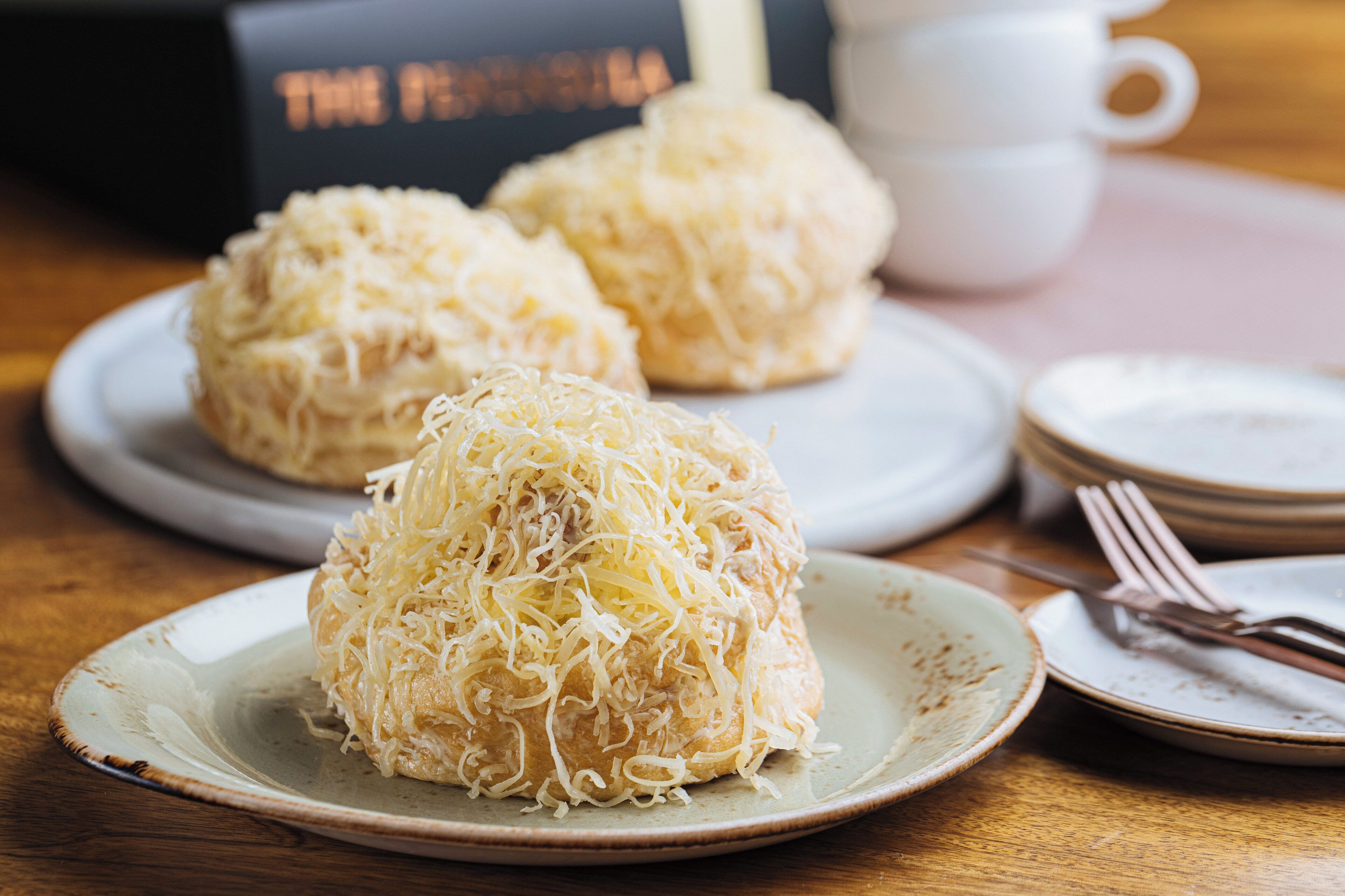 A plate with three food items covered in shredded cheese, a white cup, and a stack of plates on a wooden table.