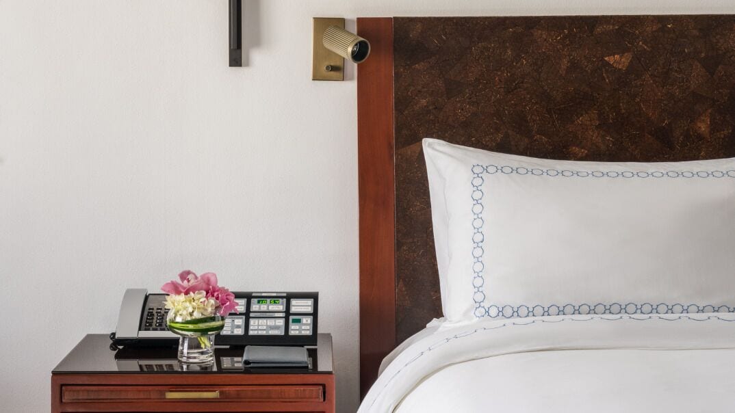 A hotel room with a bed, a nightstand with a telephone and a vase of flowers, and a wall-mounted security camera.