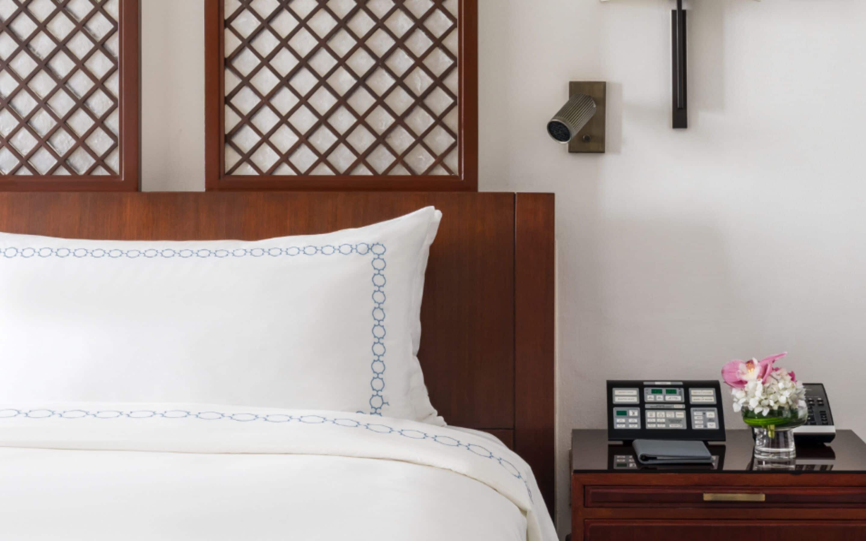 Habitación de hotel con cabecera de madera, cama blanca con diseños circulares azules, mesita de noche con un teléfono, una lámpara y un pequeño jarrón con flores.