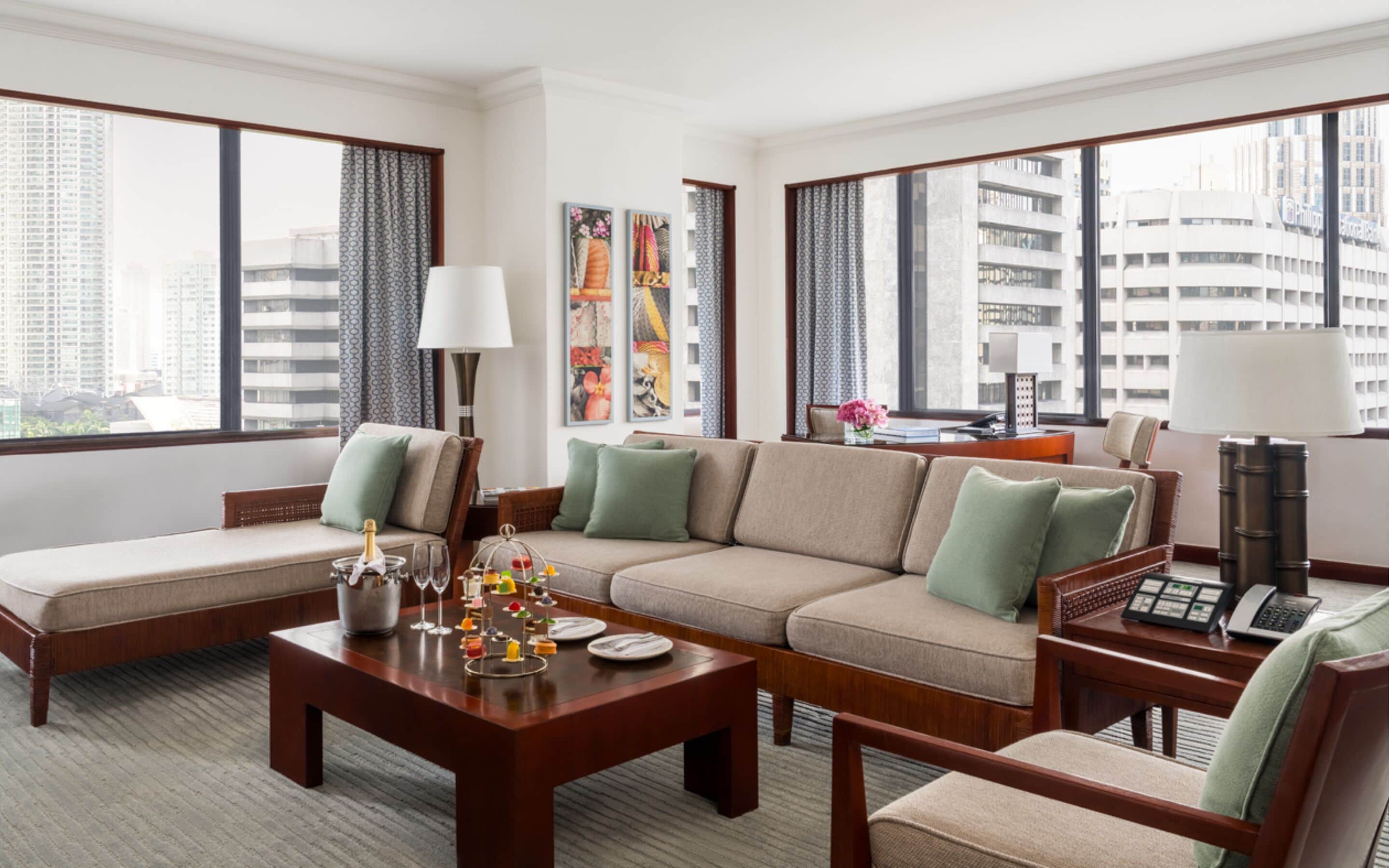 A modern hotel room with a sectional sofa, a dining table with a bottle of champagne, and a city view through large windows.