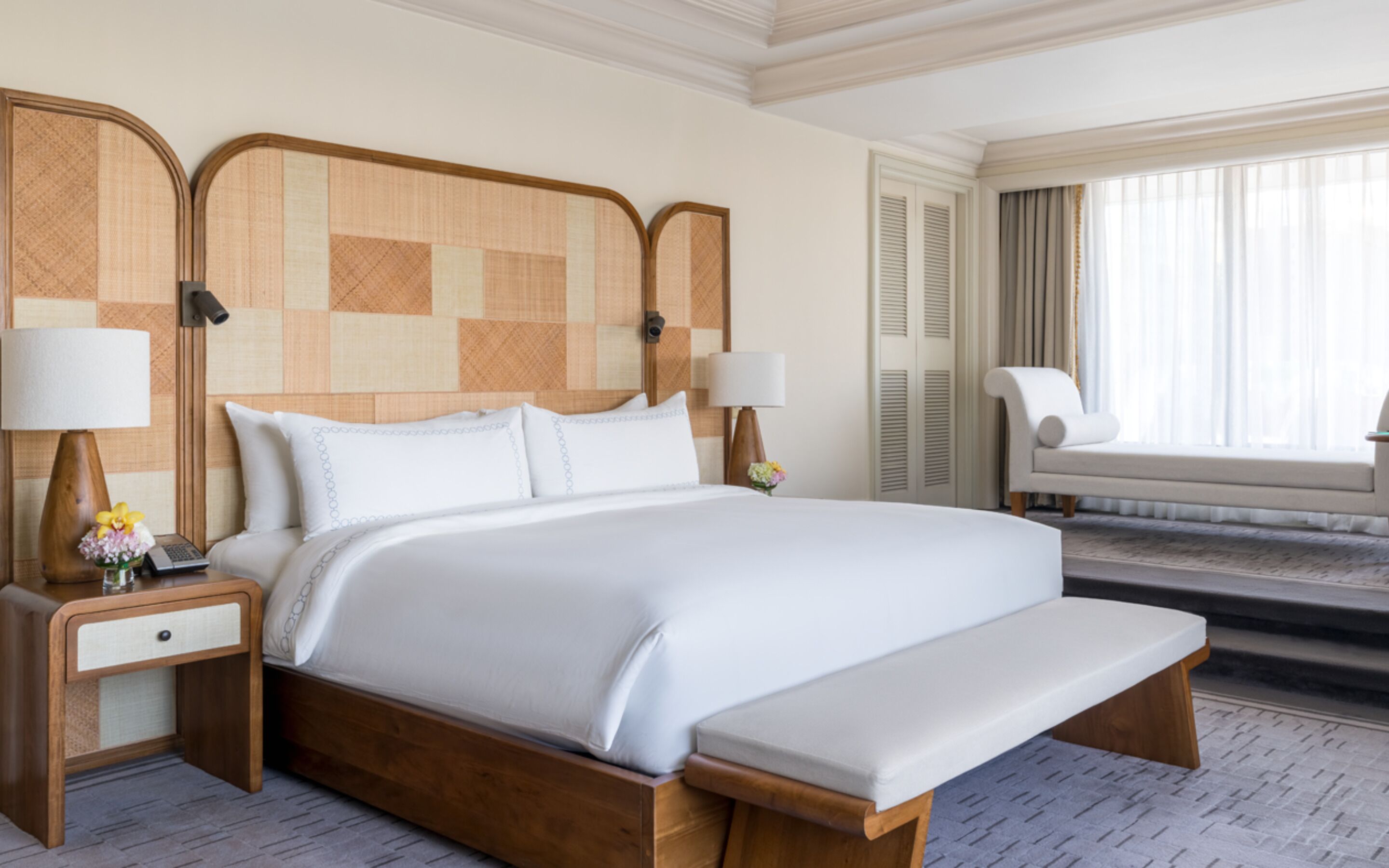 A hotel room with a large bed, two bedside tables with lamps, a bench, a chair, and a window with curtains.