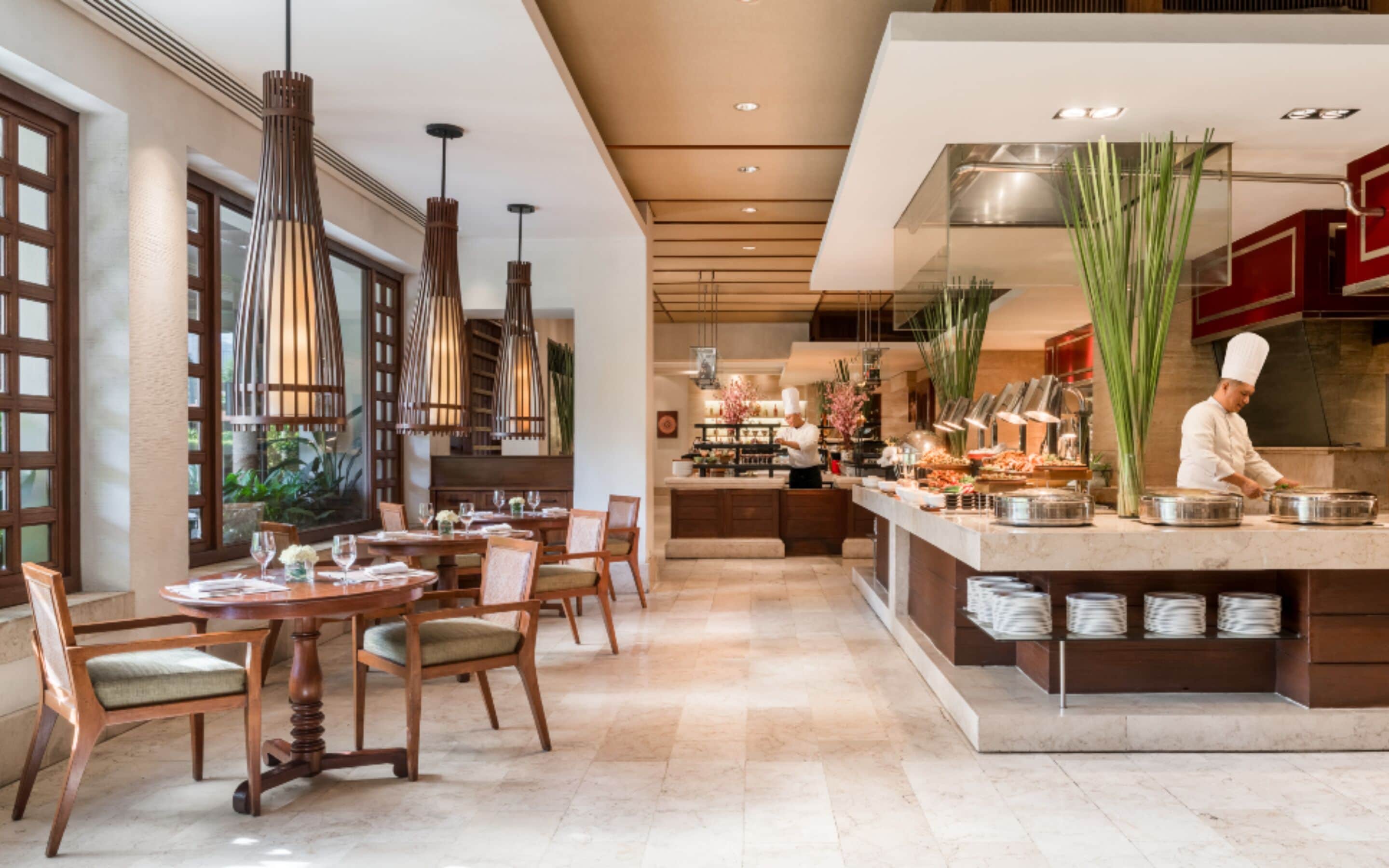 A modern restaurant with a chef preparing food, wooden furniture, and pendant lighting.