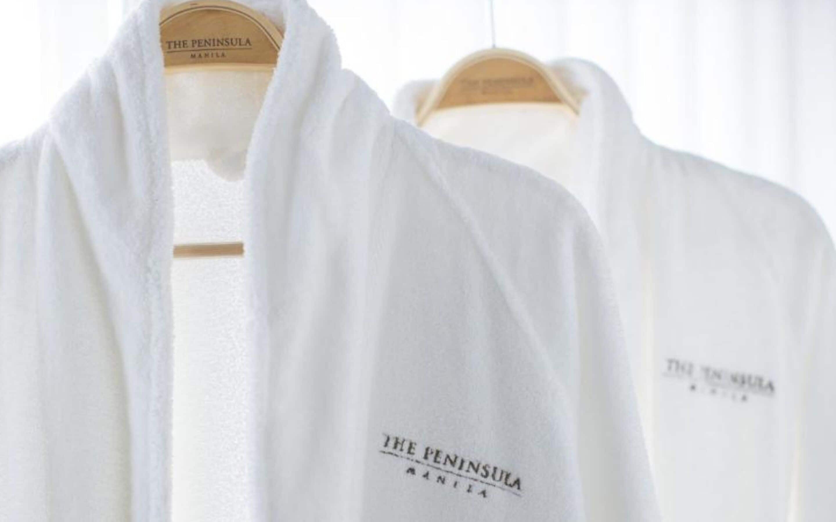 Two white robes hanging on hangers with 'THE PENINSULA MANILA' printed on them.