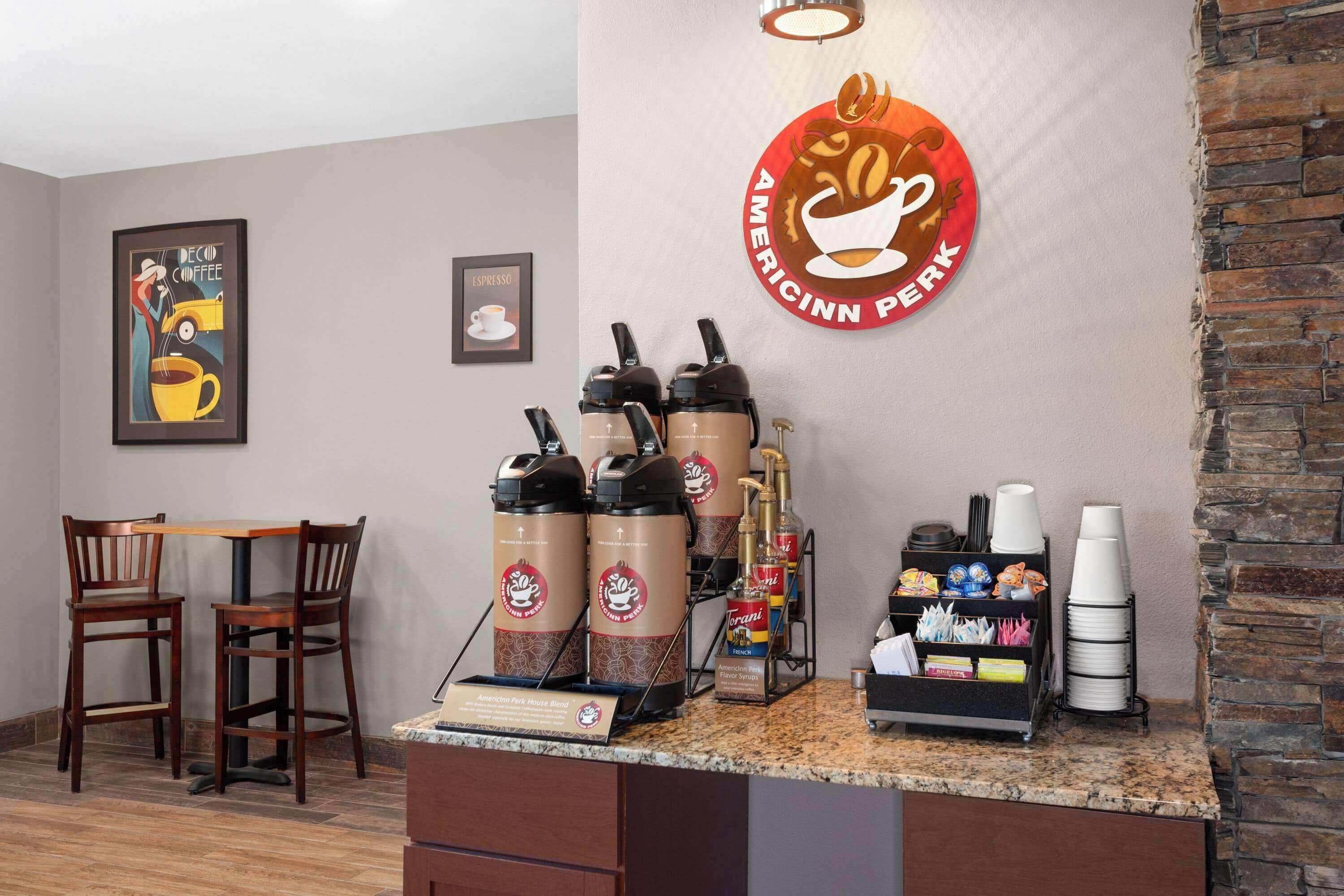 Un espace dédié au café, équipé de machines à café de la marque Americinn Perk, d’un tableau affichant le menu et d’un coin salon avec des chaises en bois.