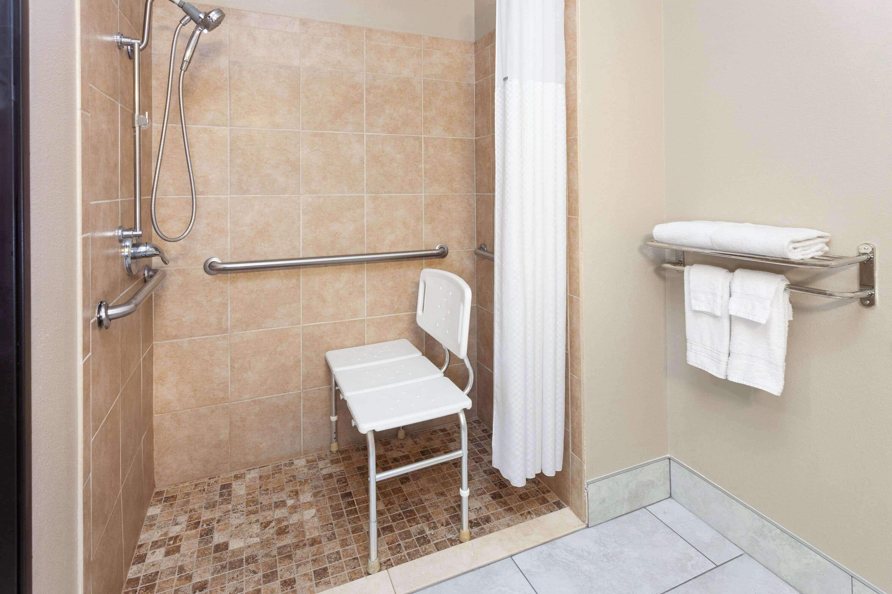 A shower area with a white chair, grab bars, a towel rack, and a showerhead.