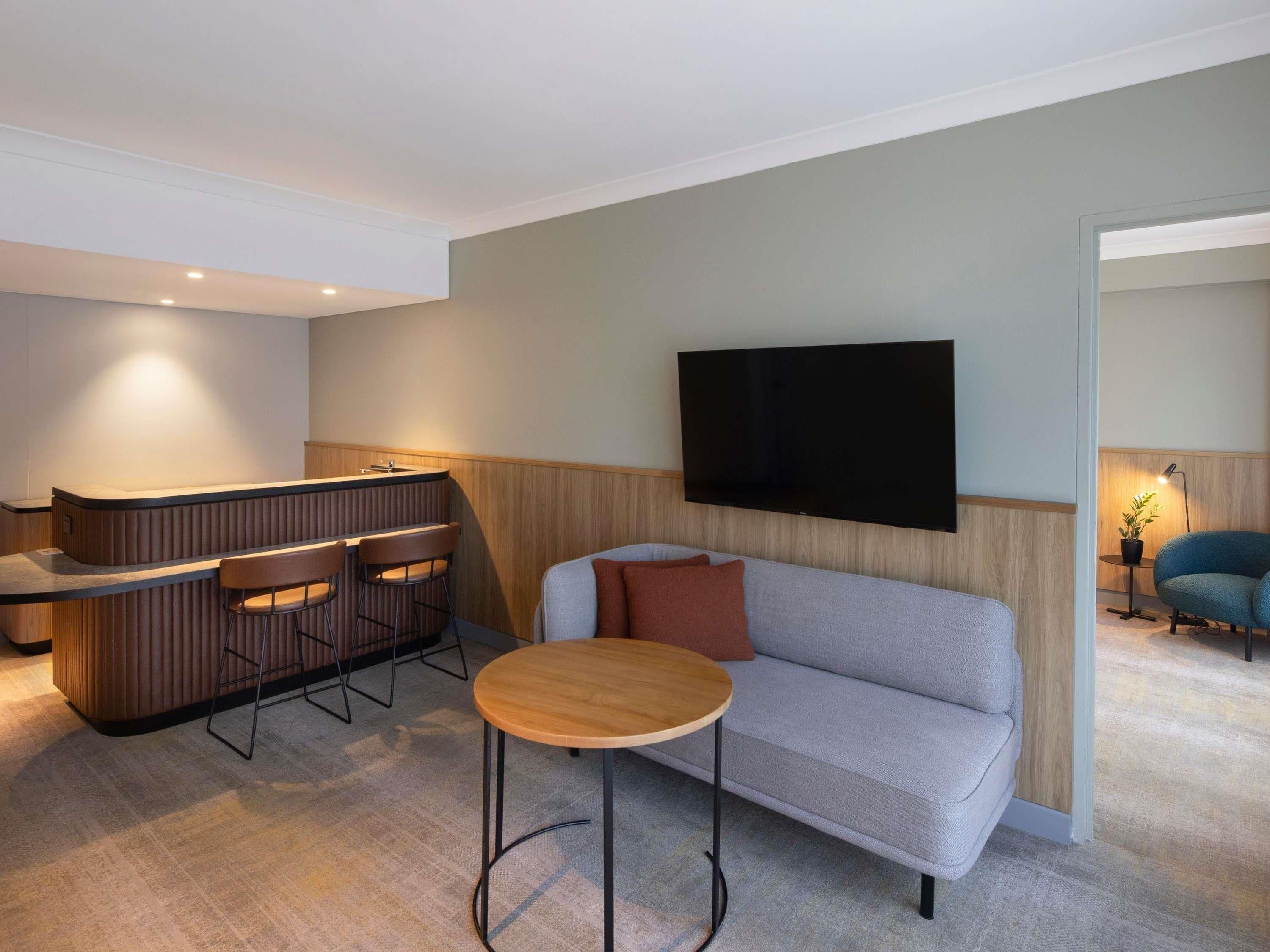 Una sala de estar moderna con un sofá, una mesa redonda de centro, un televisor montado en la pared y una zona de bar con taburetes.
