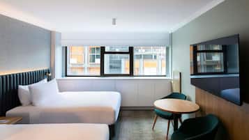 Habitación de hotel con dos camas, televisión, mesita y ventana con vistas a edificios.