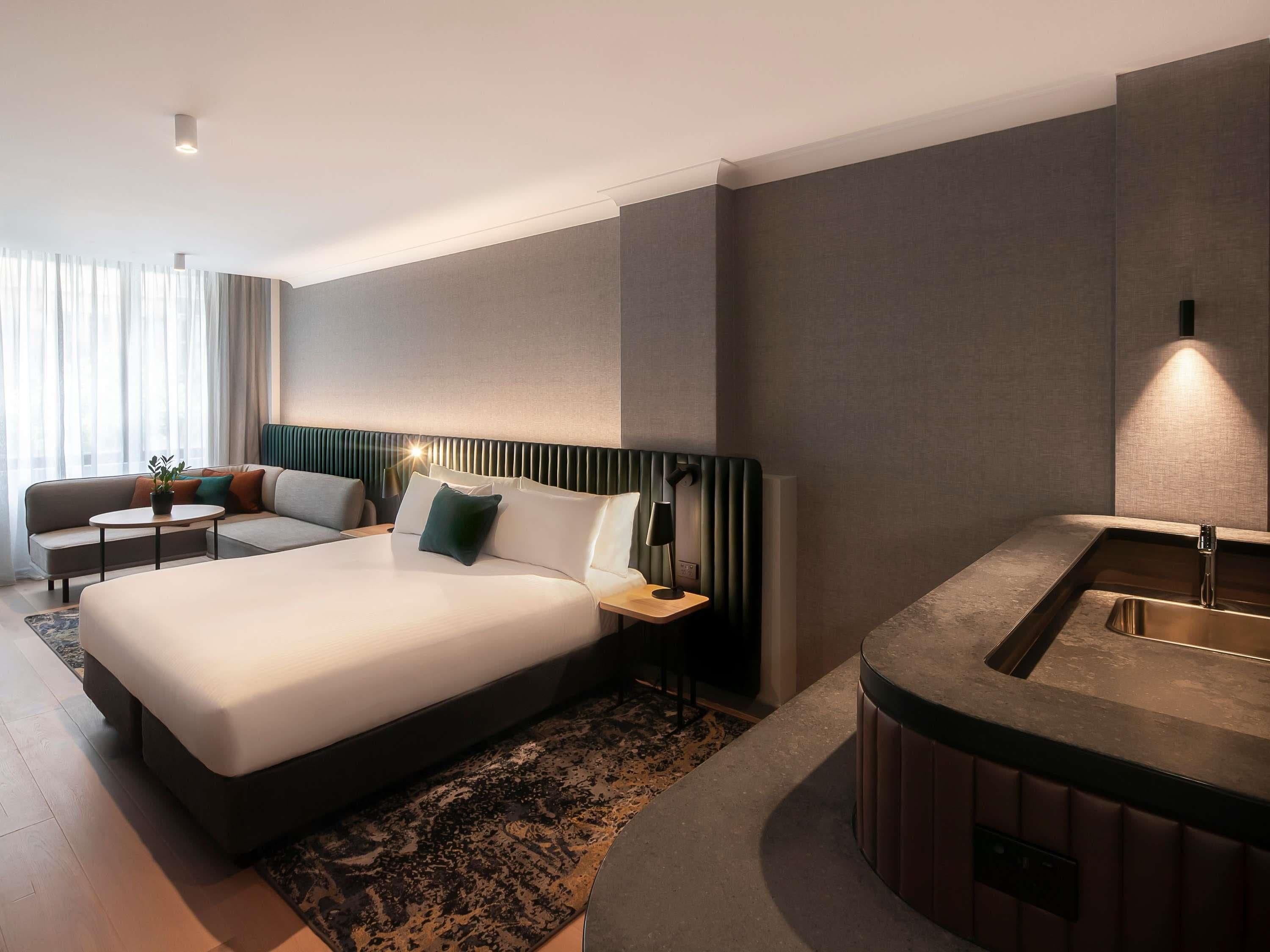 Una habitación de hotel moderna con una cama grande, un sofá, una mesita, una mesita de noche y un lavamanos integrado.