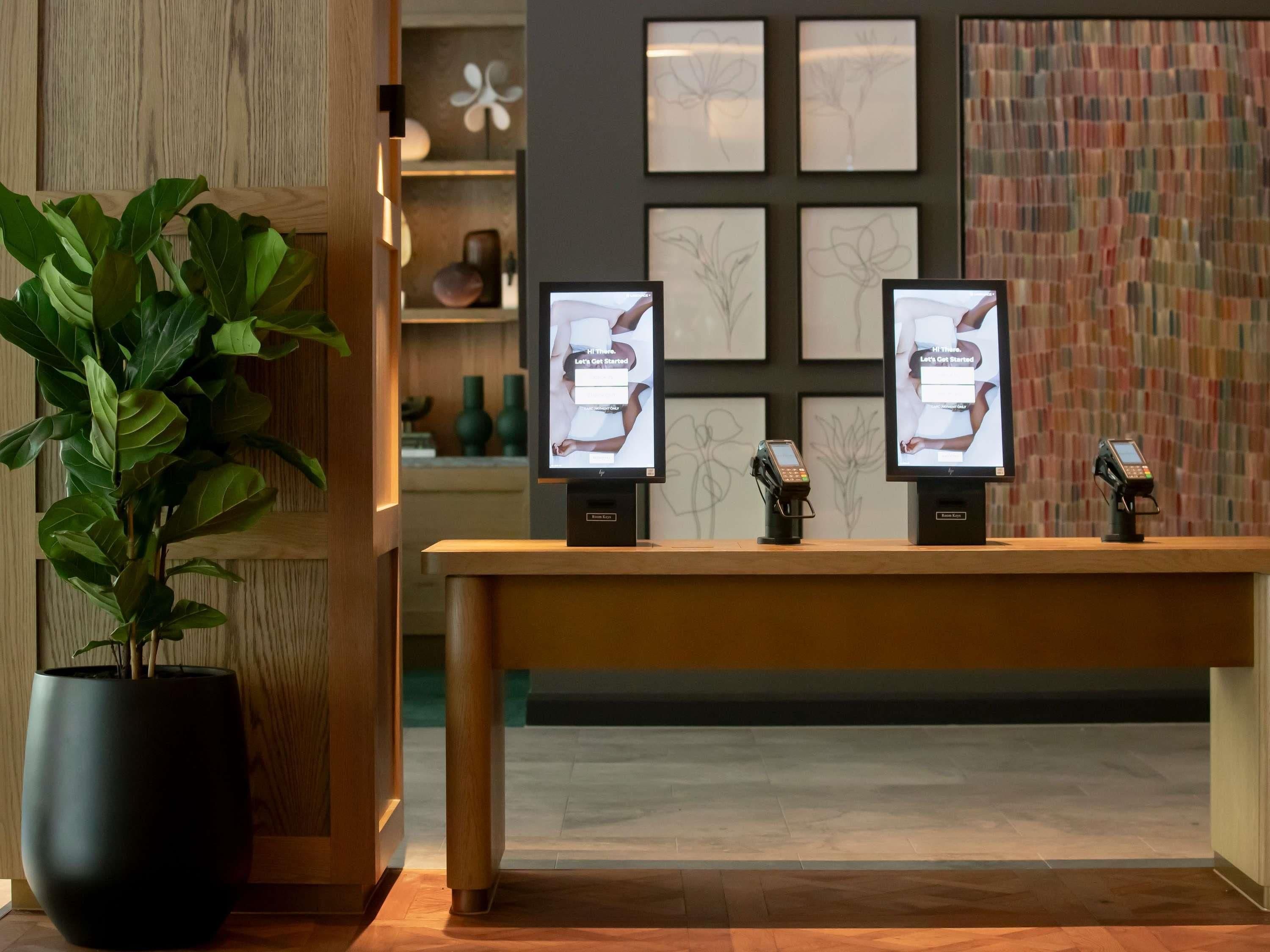 A reception desk with two monitors, a potted plant, and framed artwork on the wall.