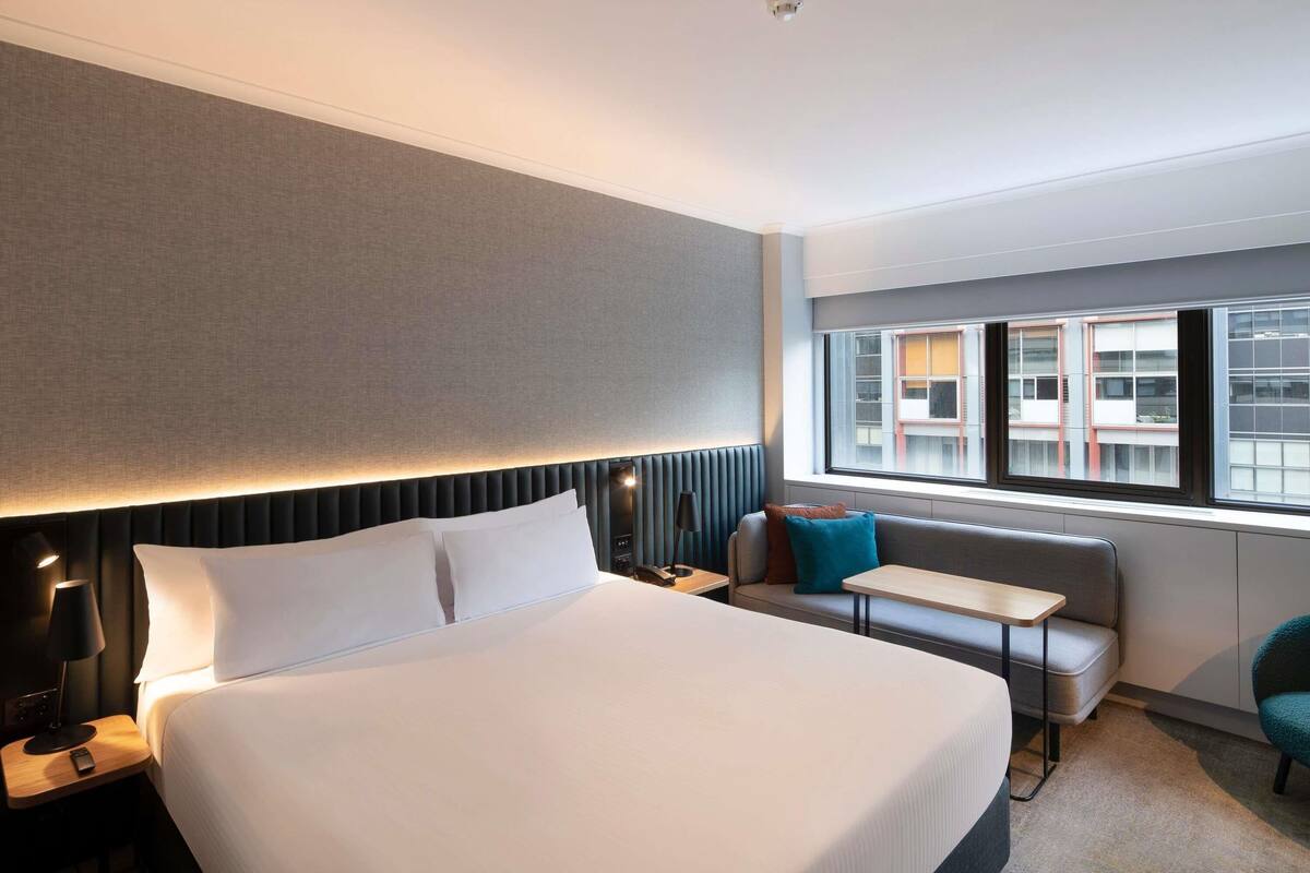 A hotel room with a large bed, a bedside table, a small table, a sofa, and a window with a view of buildings.