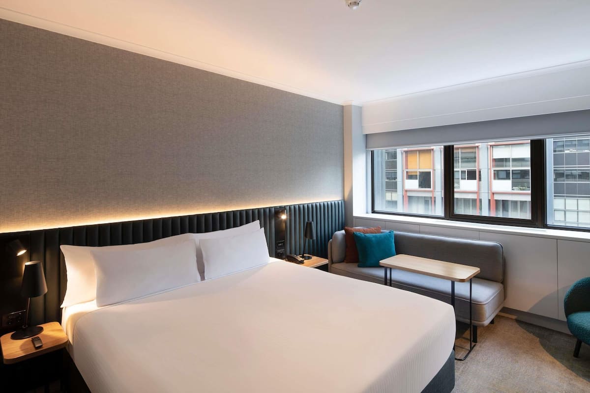A hotel room with a large bed, a bedside table, a small table, a sofa, and a window with a view of buildings.