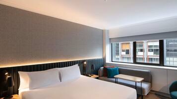 A hotel room with a large bed, a bedside table, a small table, a sofa, and a window with a view of buildings.