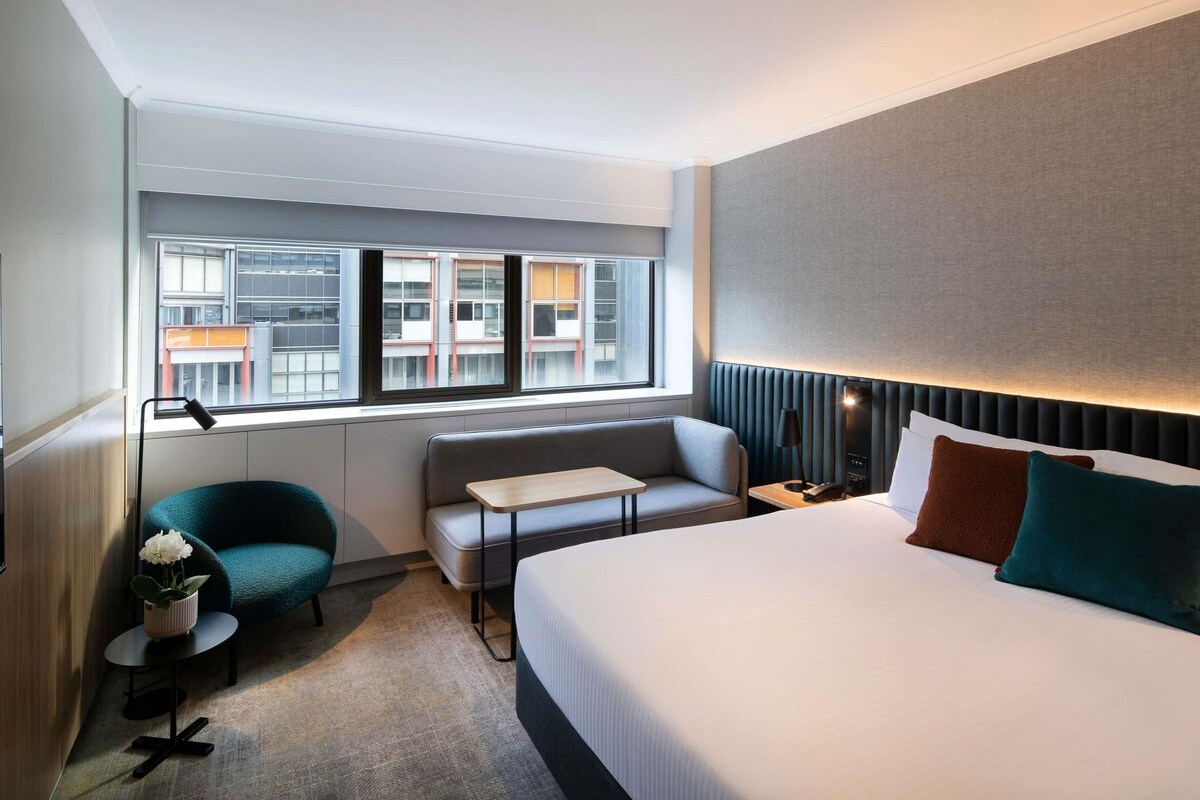 A hotel room with a bed, a sofa, a small table, a lamp, a potted plant, and a view of buildings through the window.