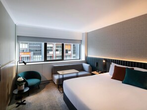 Habitación de hotel con cama, sofá, una mesita, lámpara, planta en maceta y vistas de edificios a través de la ventana.