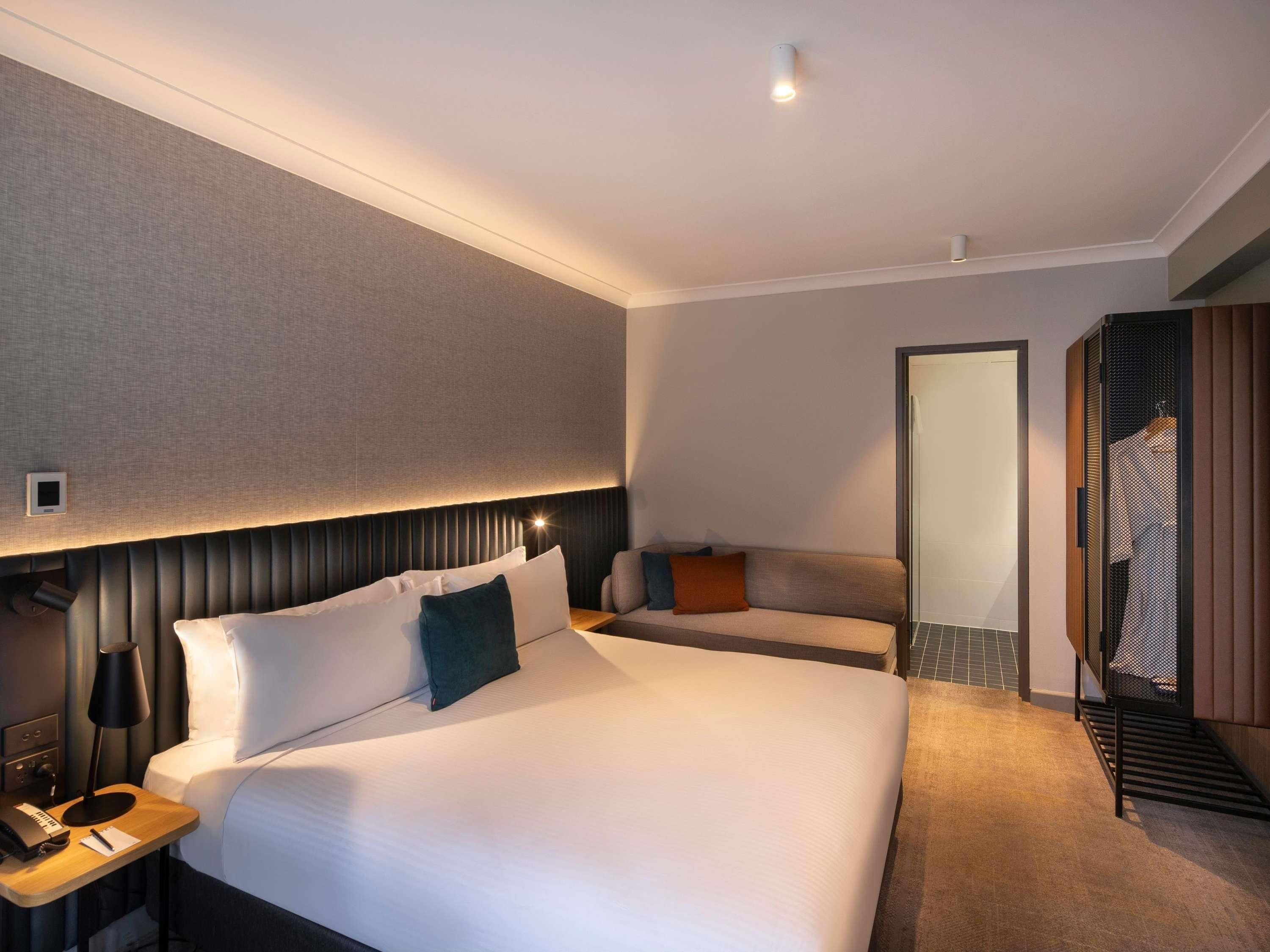 A hotel room with a large bed, a bedside table with a lamp, a small desk with a phone and a notepad, a sofa, and a mirror.