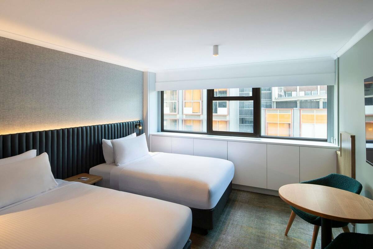 A hotel room with two beds, a small table, and a window with a view of buildings.