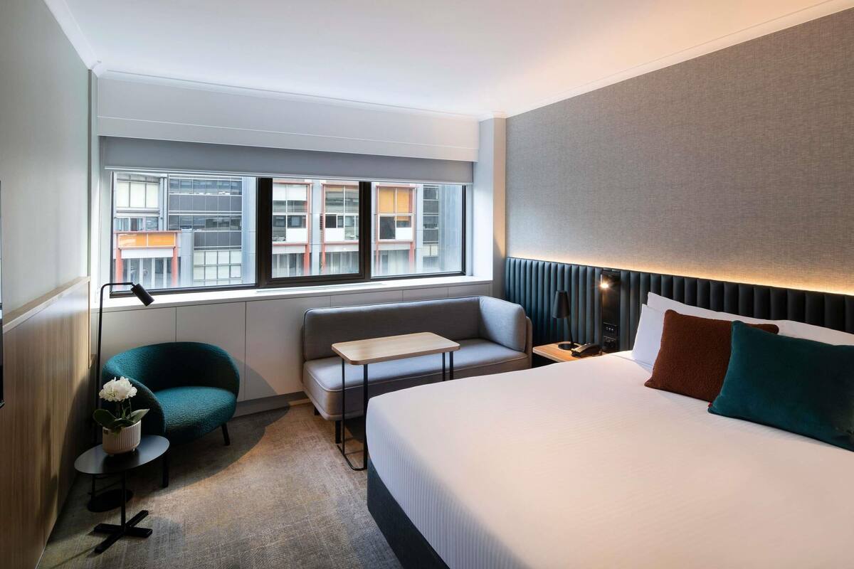 A hotel room with a bed, a sofa, a small table, a lamp, a potted plant, and a view of buildings through the window.