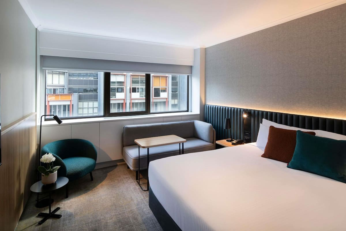 A hotel room with a bed, a sofa, a small table, a lamp, a potted plant, and a view of buildings through the window.