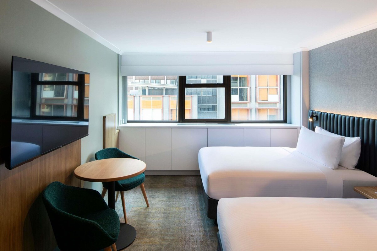 A hotel room with a bed, a TV, a small table, and a window with a city view.