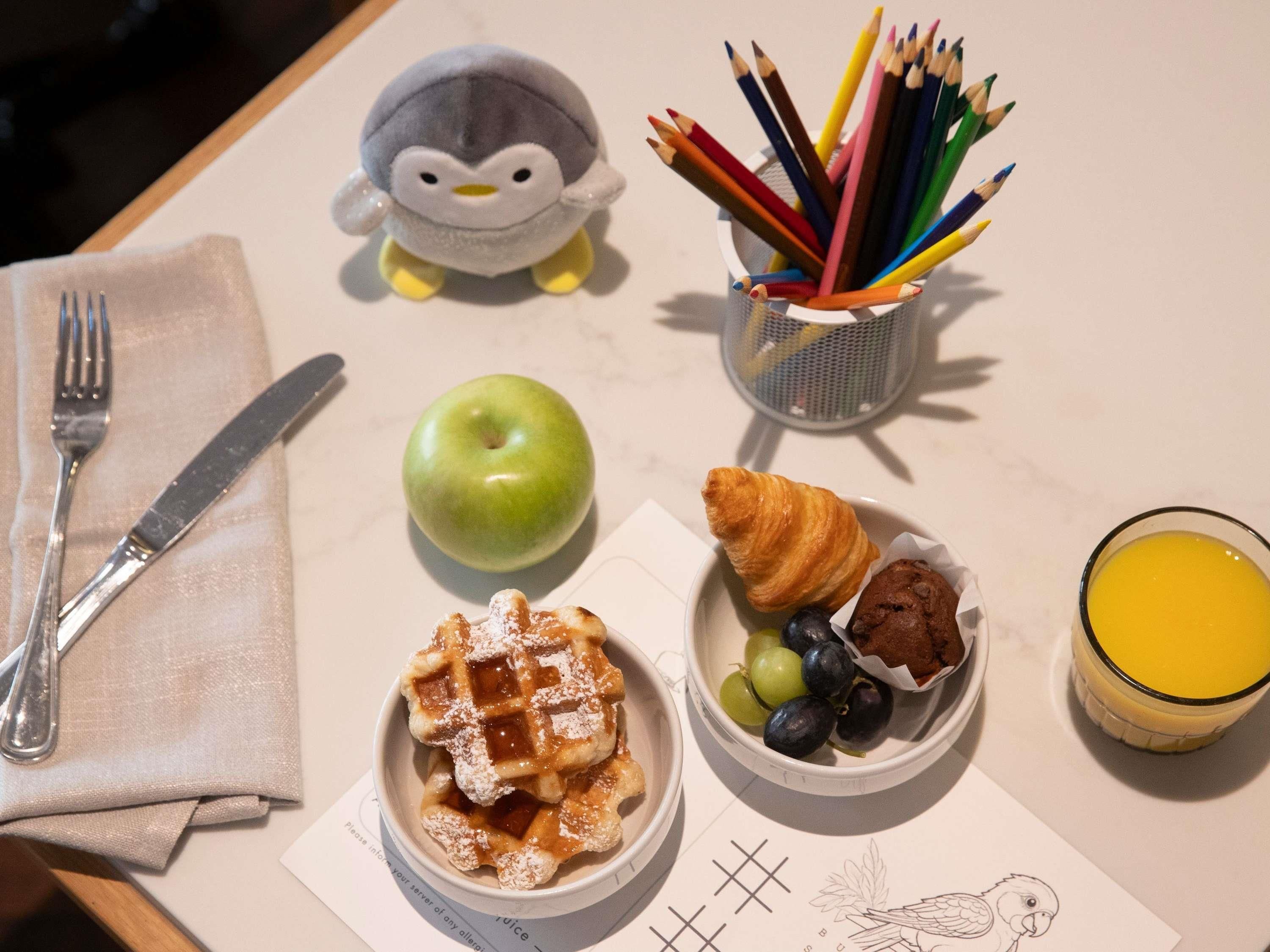 A table with a fork, knife, a stuffed penguin, a basket of colored pencils, a green apple, waffles with powdered sugar, a croissant with grapes and chocolate, and a glass of orange juice.