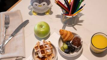 A table with a fork, knife, a stuffed penguin, a basket of colored pencils, a green apple, waffles with powdered sugar, a croissant with grapes and chocolate, and a glass of orange juice.