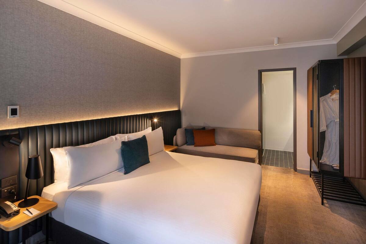A hotel room with a large bed, a bedside table with a lamp, a small desk with a phone and a notepad, a sofa, and a mirror.