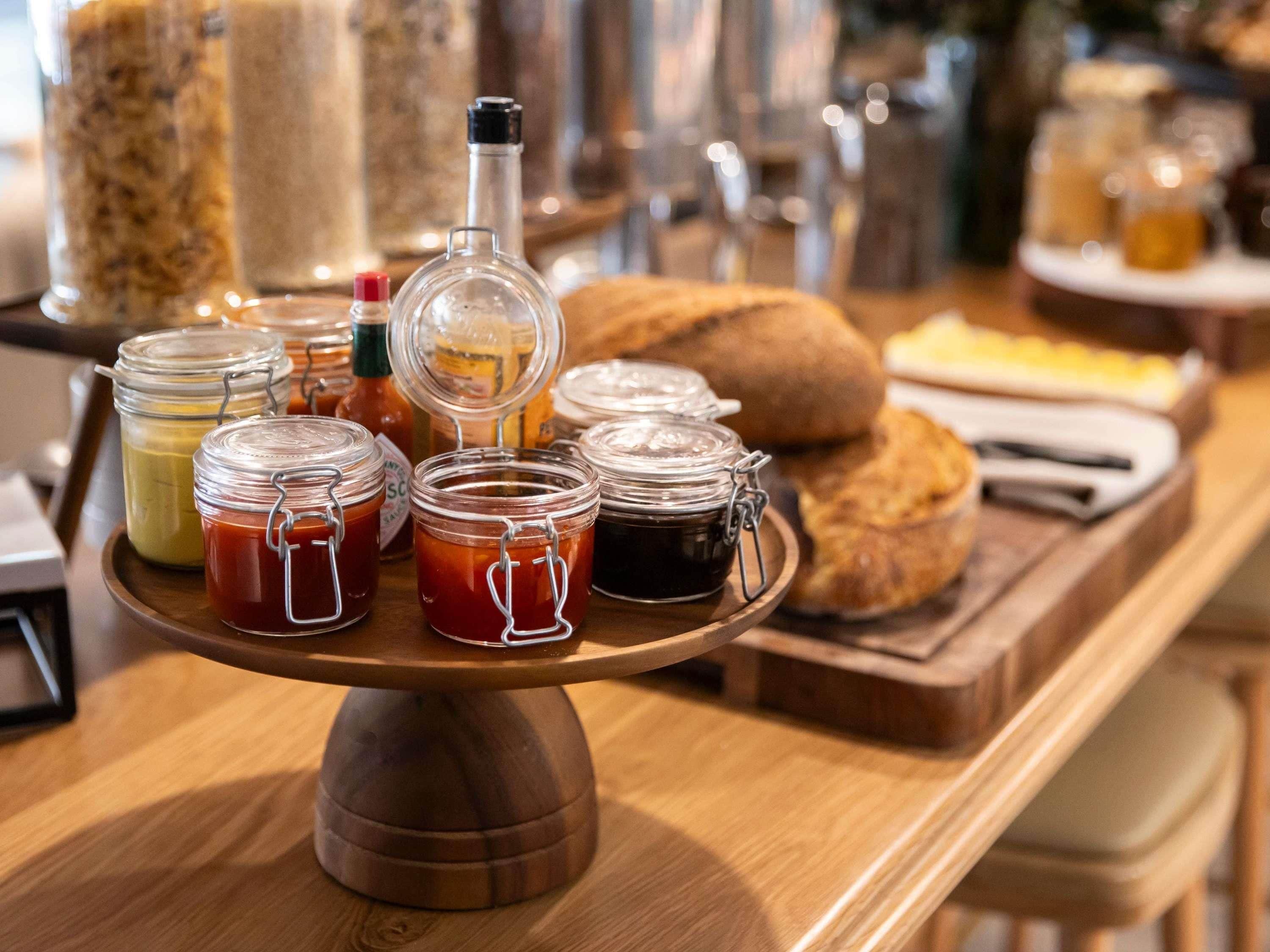 A wooden serving tray with jars of condiments and a bottle on a table.