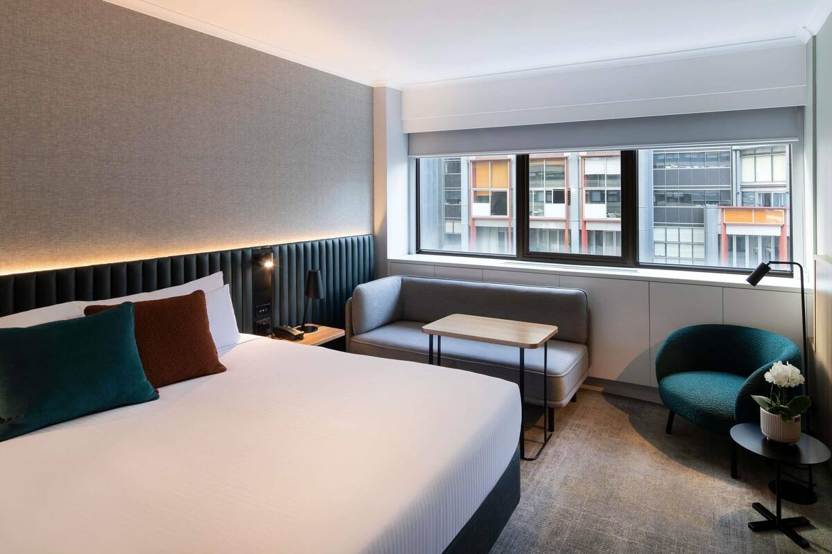 A hotel room with a bed, a sofa, a small table, a chair, a nightstand, a lamp, and a window with a city view.