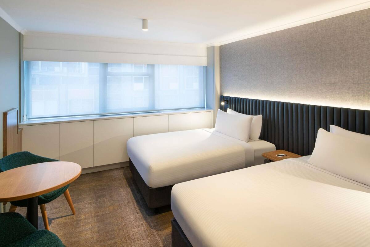 A hotel room with two beds, a small table, and a window with blinds.