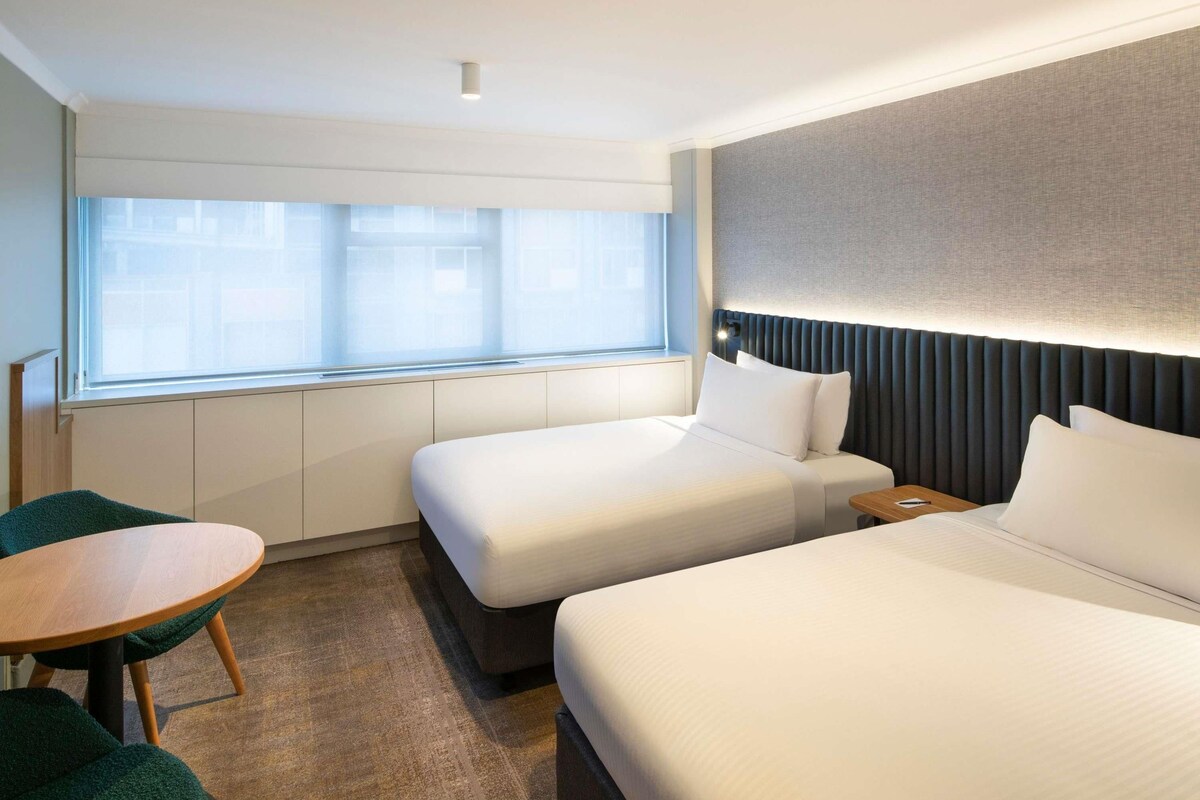 A hotel room with two beds, a small table, and a window with blinds.