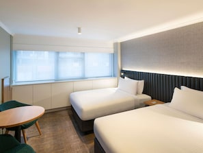 A hotel room with two beds, a small table, and a window with blinds.