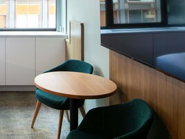 A small, round wooden table with two dark green chairs, a window with blinds, and a view of buildings outside.