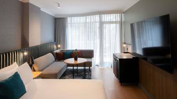 Habitación de hotel con cama, sofá, una mesita, televisor y ventana con cortinas.