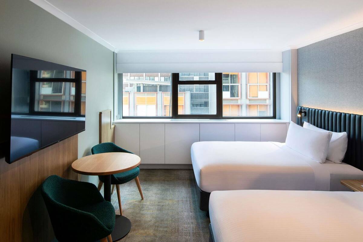 A hotel room with a bed, a TV, a small table, and a window with a city view.