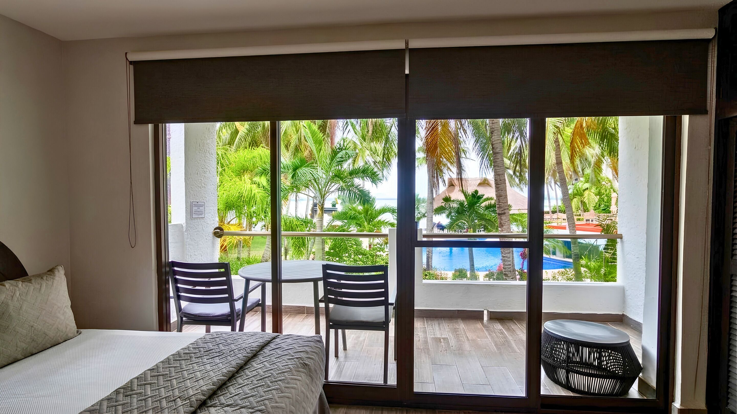 A hotel room with a bed, a table, two chairs, a small round table, and a view of a pool and palm trees through large sliding glass doors.