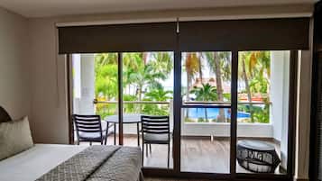 Habitación de hotel con cama, una mesa, dos sillas, una mesita redonda y vista a una piscina y palmeras a través de puertas correderas de vidrio.