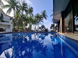 Piscina com reflexos de palmeiras, rodeada por espreguiçadeiras e fachada de um edifício.
