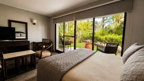 A bedroom with a large bed, a desk, a chair, and a television. There is a window with a view of greenery outside. - Sina Suites Cancún - Adults Only (Cancun)