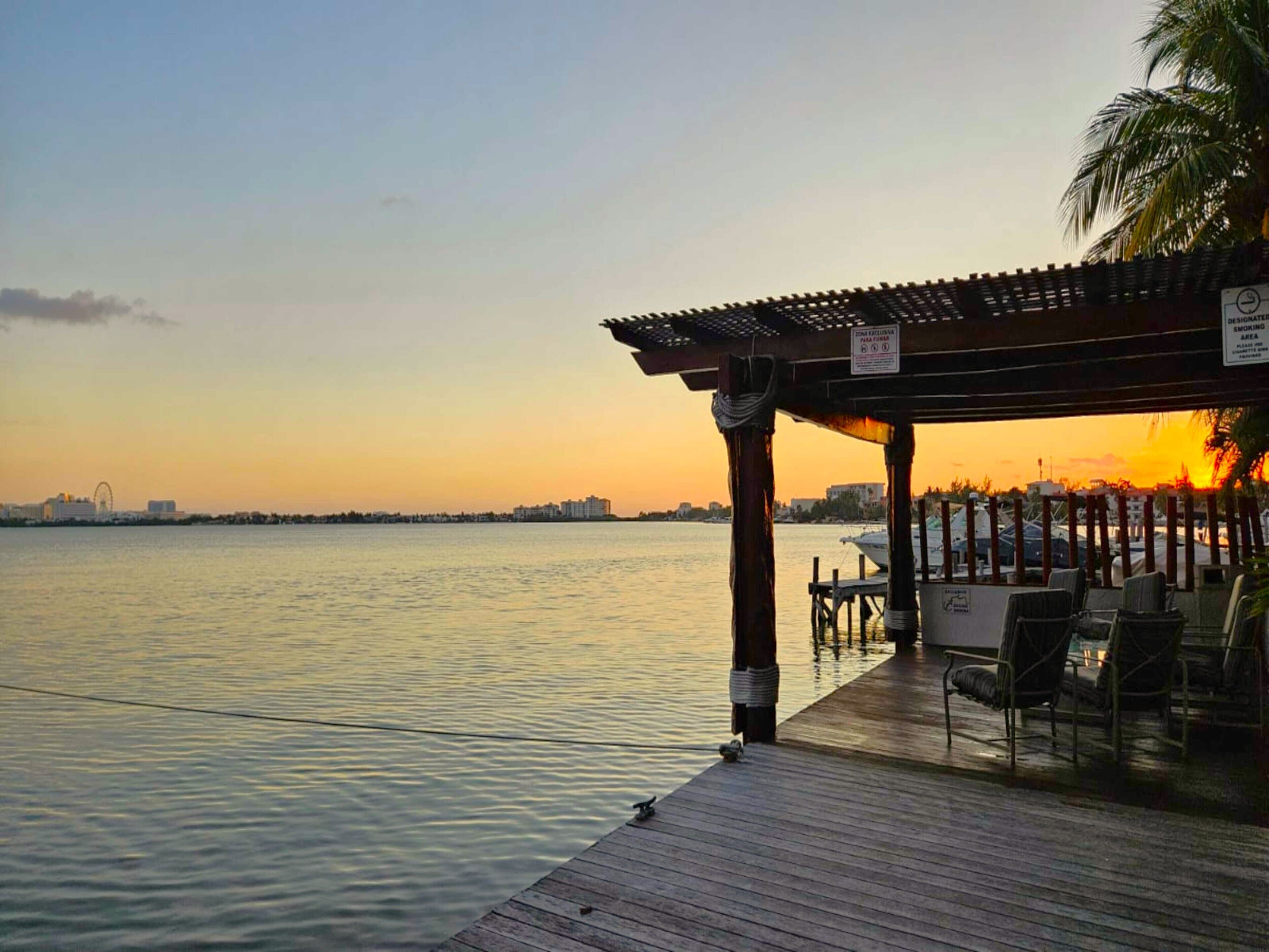 A wooden pier extending into calm waters at sunset, with a covered seating area and a palm tree on the right.