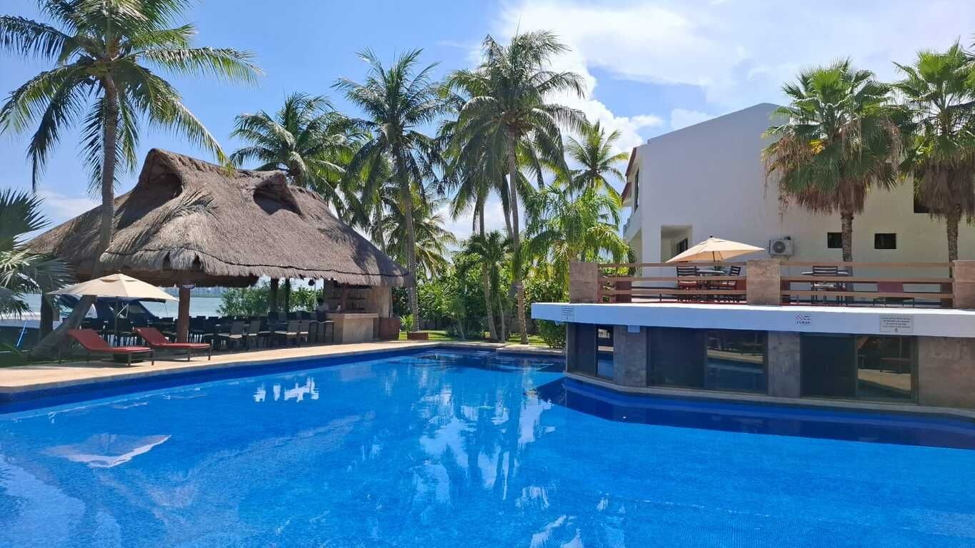 A swimming pool with lounge chairs and umbrellas, surrounded by palm trees and a building with a white facade.