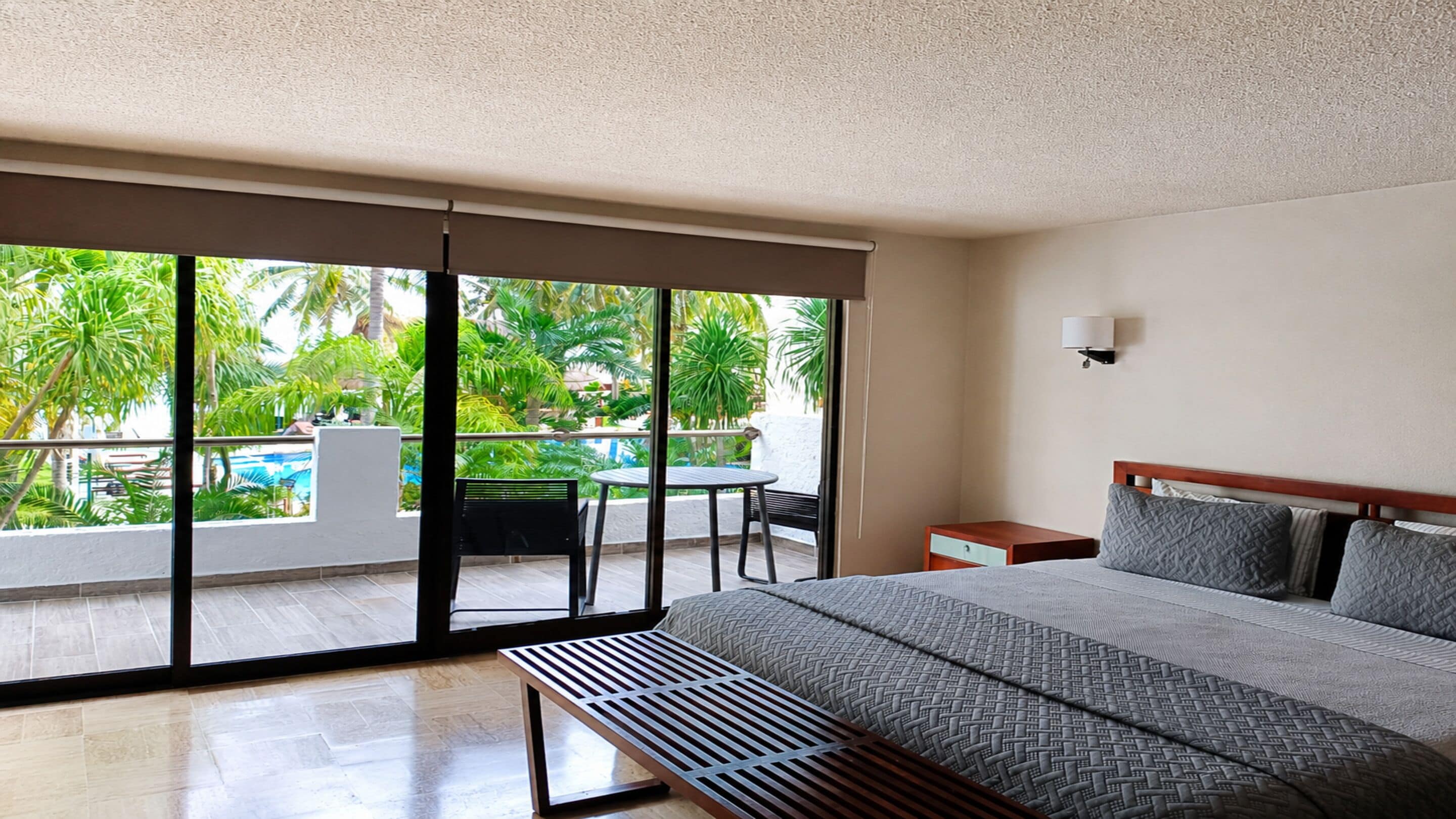 A hotel room with a large bed, a small table, a bench, and a view of a pool and palm trees through the balcony doors.