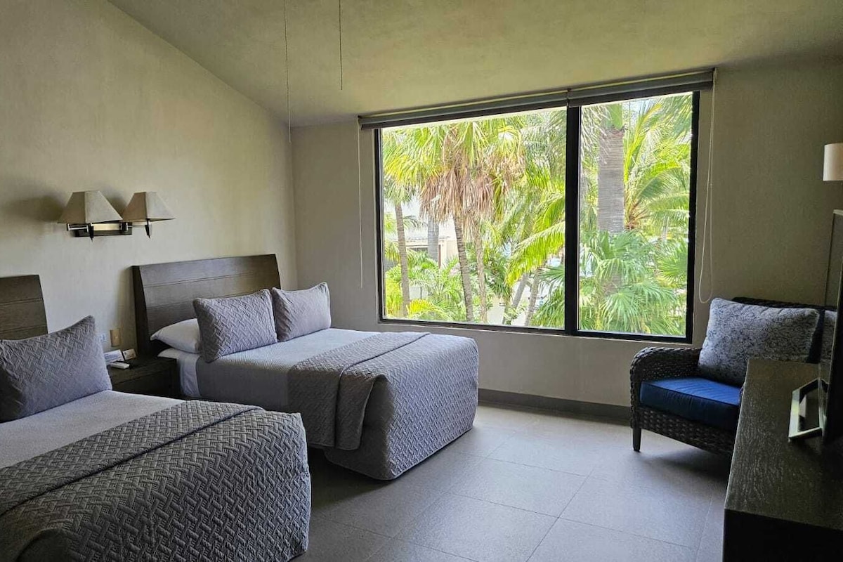 Habitación de hotel con dos camas, una mesita de noche, una silla y un amplio ventanal con vistas a palmeras.