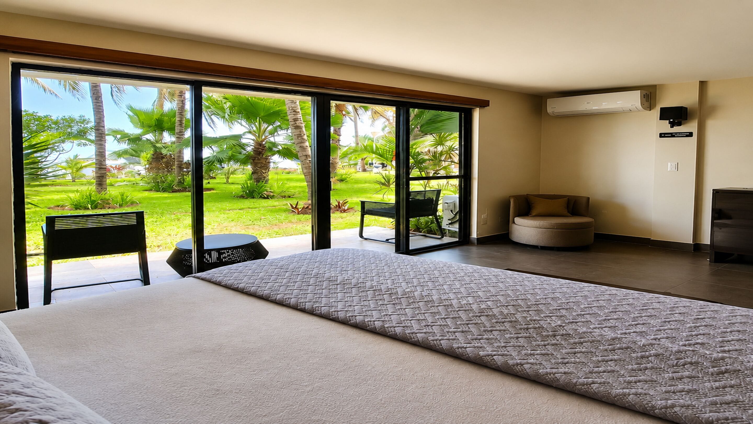 Habitación de hotel con una cama grande, una mesita pequeña, dos sillas y vistas a un jardín tropical a través de puertas correderas amplias.