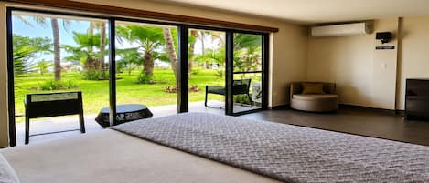 Habitación de hotel con una cama grande, una mesita pequeña, dos sillas y vistas a un jardín tropical a través de puertas correderas amplias.