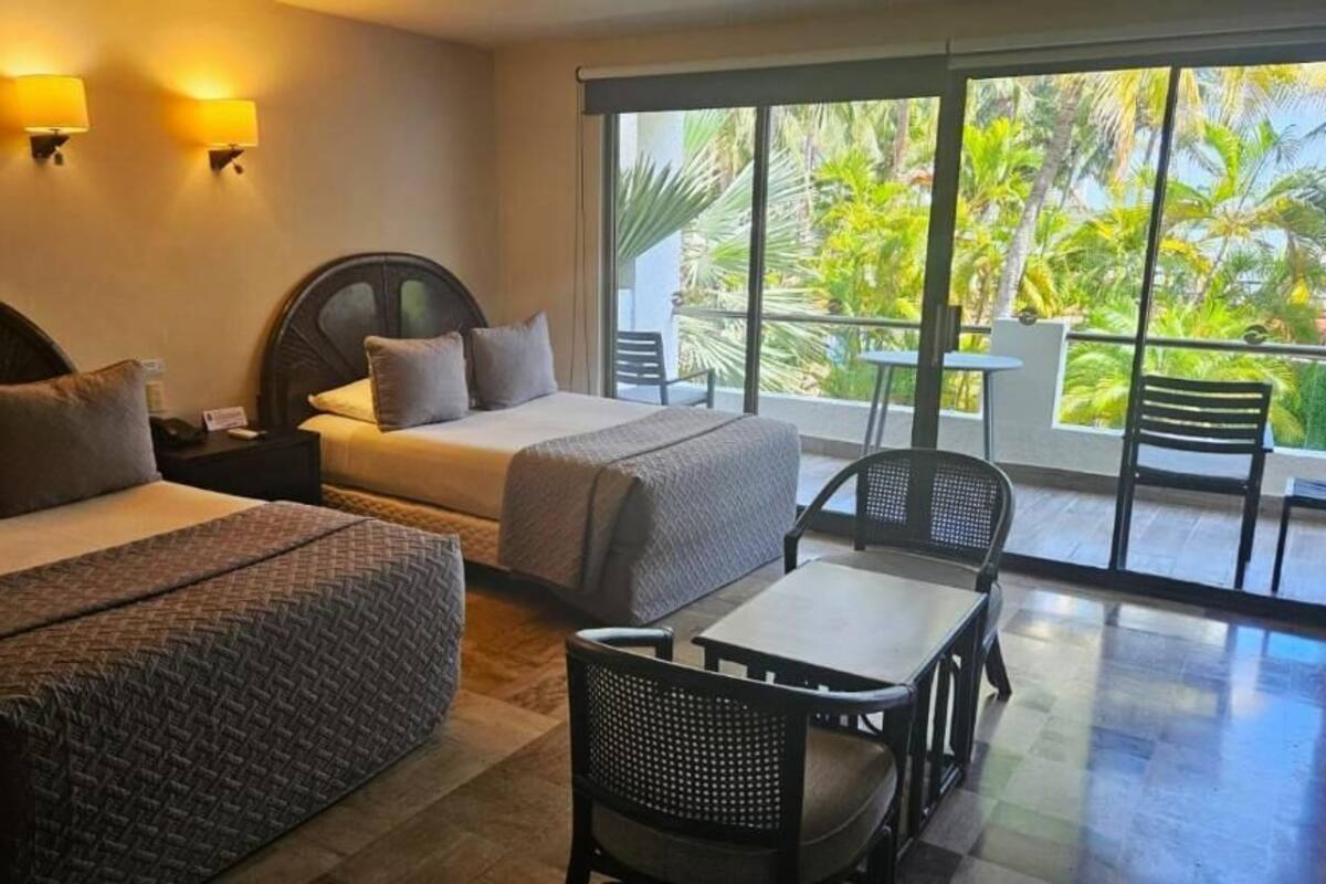 A hotel room with two beds, a balcony with a table and chairs, and a view of palm trees.