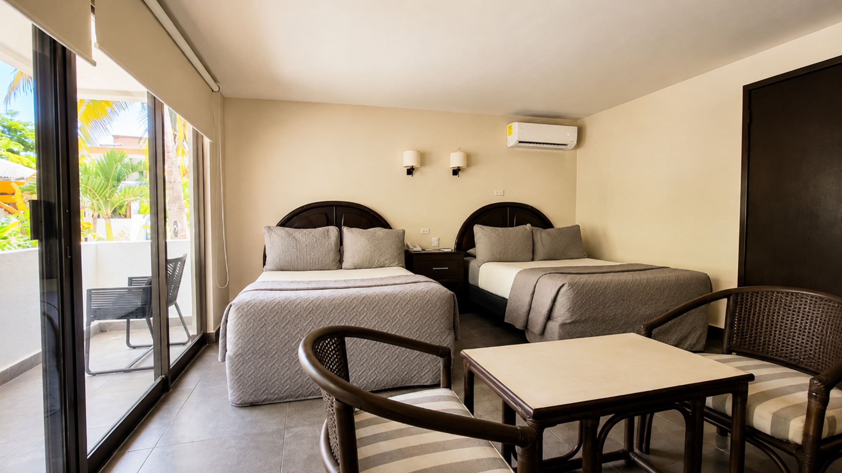 A hotel room with two beds, a small table, two chairs, a balcony with a chair, and an air conditioning unit.