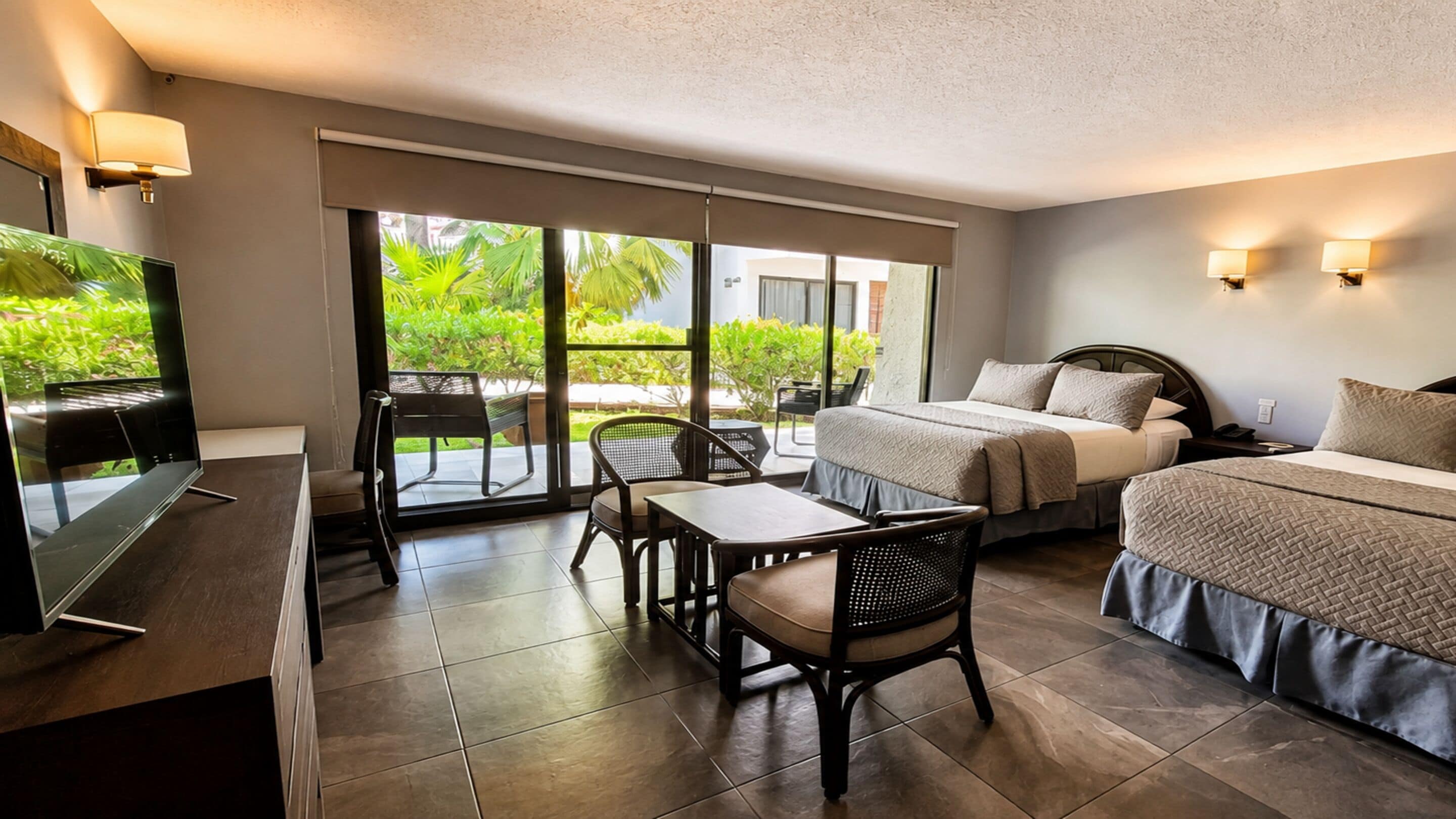 A hotel room with a bed, a desk, a chair, a TV, and a sliding glass door leading to a balcony with greenery.